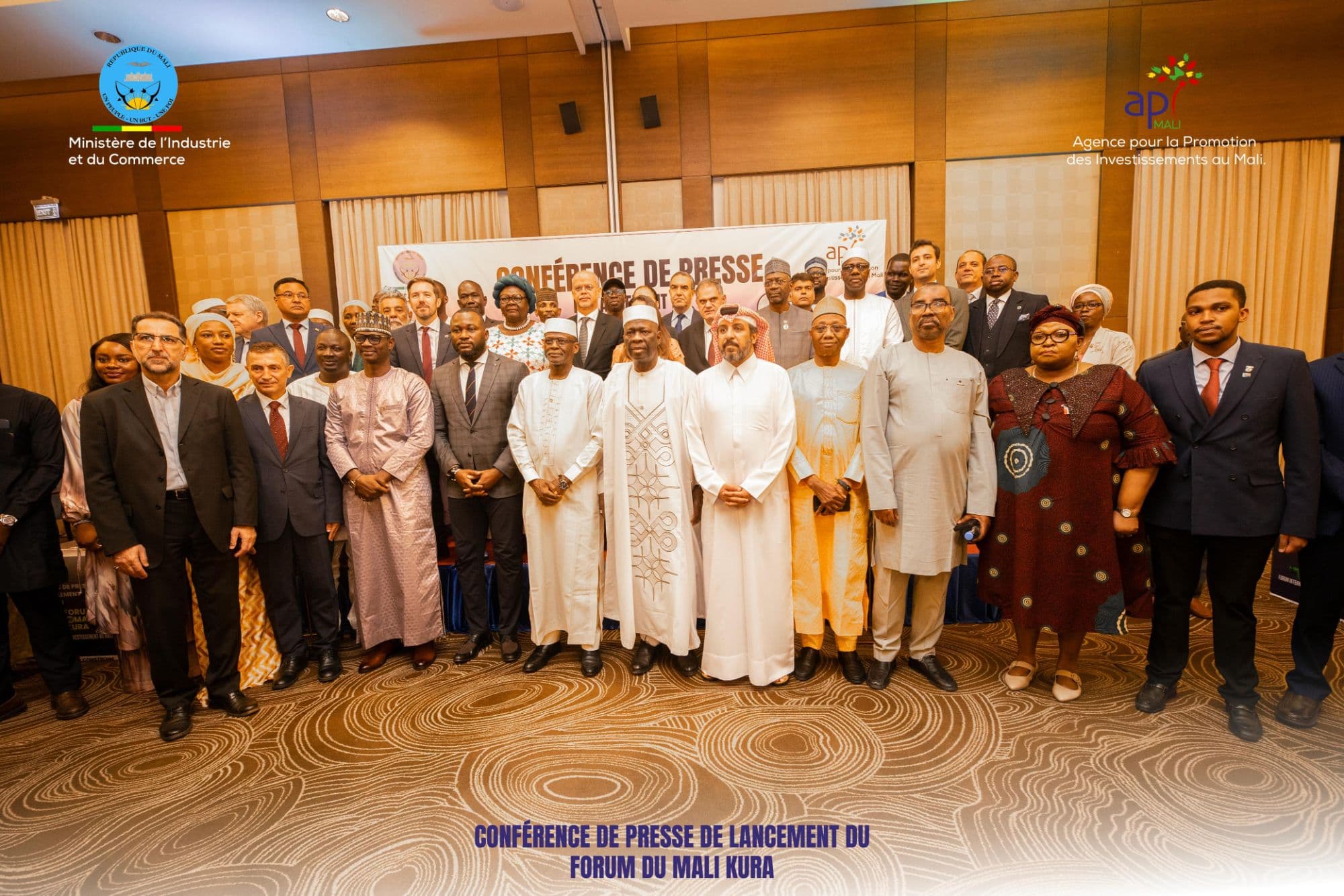 Group photo of the Mali Kura Forum launch press conference: some fifty officials, diplomats and investors in suits, grand boubous and Gulf dress, standing in two rows in front of the “Conférence de presse” banner, with the logos of the Ministry of Industry and Trade and of API-Mali at the top of the frame. — API-Mali