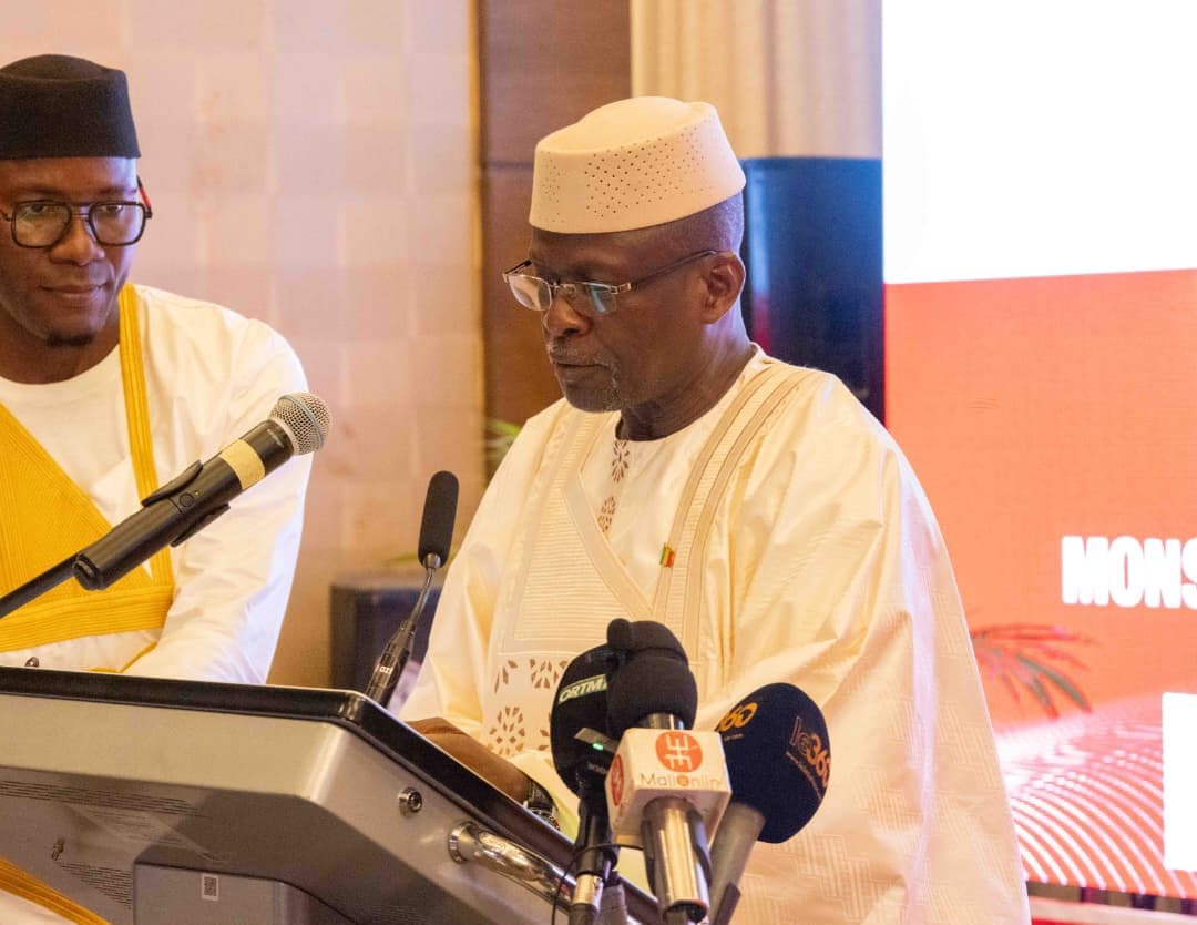 A man in a cream boubou and matching cap reads his speech at a lectern fitted with media microphones, another man in a white and yellow boubou beside him, a red screen behind.