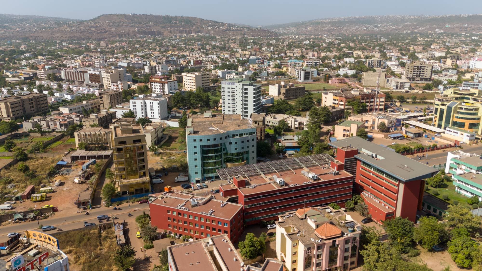 Aerial view of a Bamako business district: a large red office building with solar panels on its roof, a blue-green glass tower, buildings under construction, an avenue with motorcycles and cars, and the city and hills on the horizon.