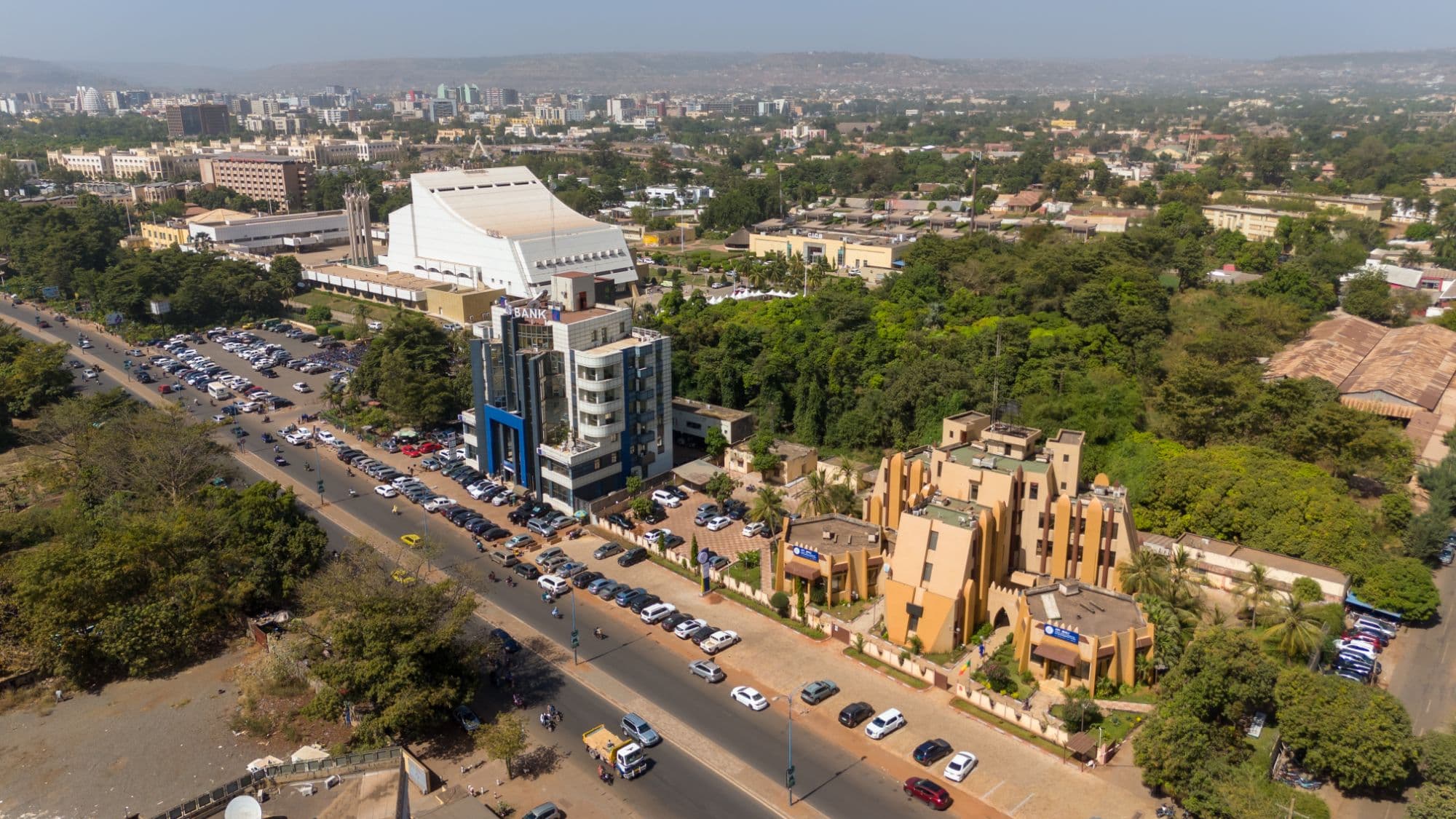 Vue aérienne d’ACI 2000 à Bamako : le Centre international de conférences, grand bâtiment blanc à la toiture inclinée, au-dessus d’un boulevard bordé de voitures garées, d’un immeuble de bureaux bleu et blanc et d’un bâtiment ocre de style soudanais, au milieu d’une large masse d’arbres, la ville et les collines à l’horizon.