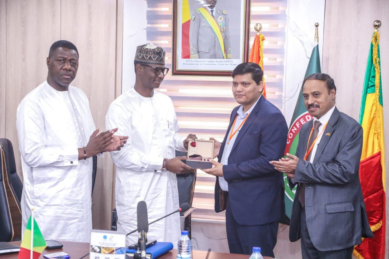 Remise d’un coffret lors d’une rencontre officielle : deux hommes en grand boubou blanc et deux hommes en costume, cordon de badge orange au cou, devant le portrait officiel du chef de l’État et les drapeaux du Mali et de la Confédération des États du Sahel.