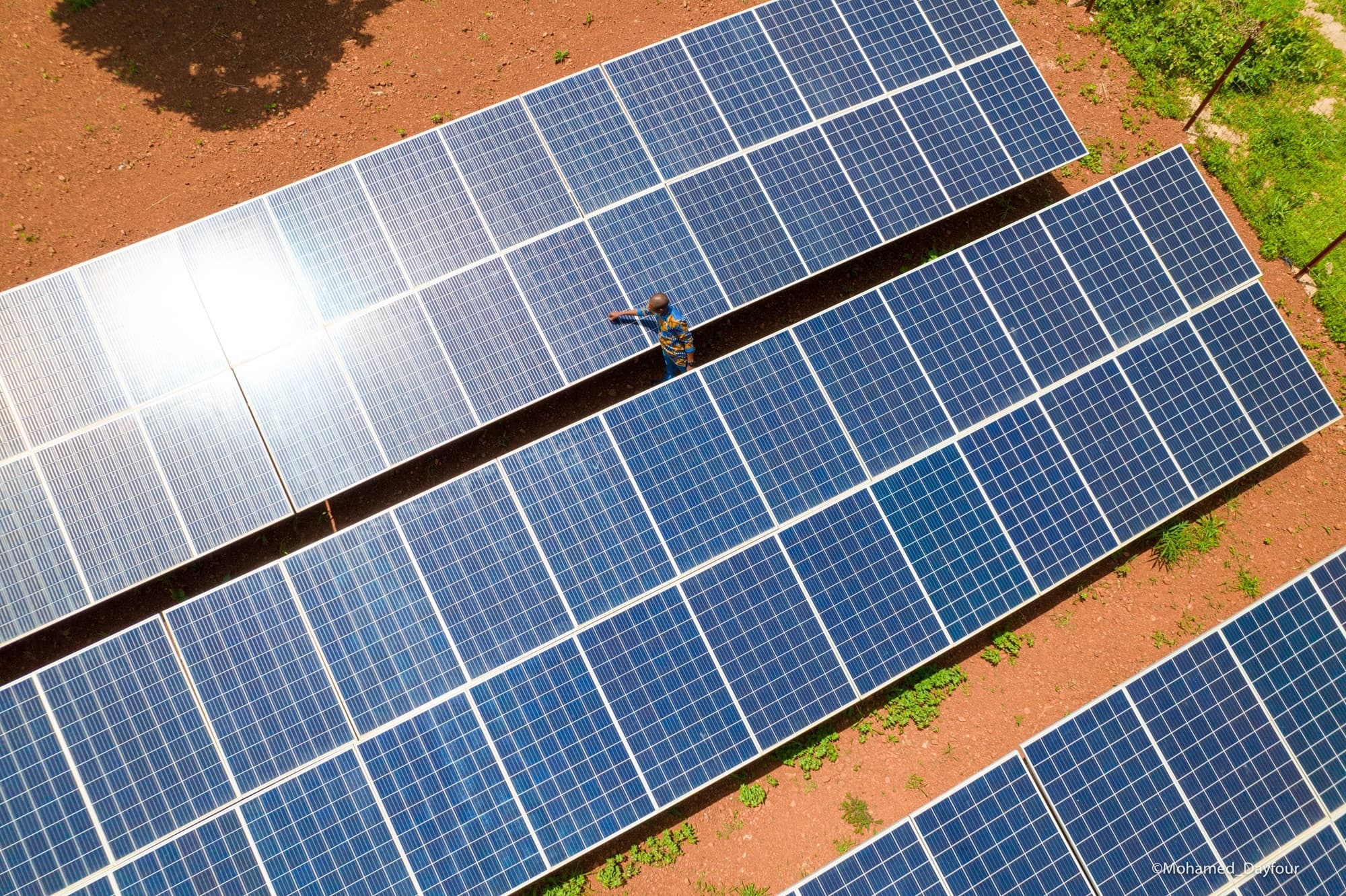 Overhead view of three long rows of blue photovoltaic panels on red laterite soil, with a technician cleaning a panel between two rows. — Mohamed Dayfour / API-Mali