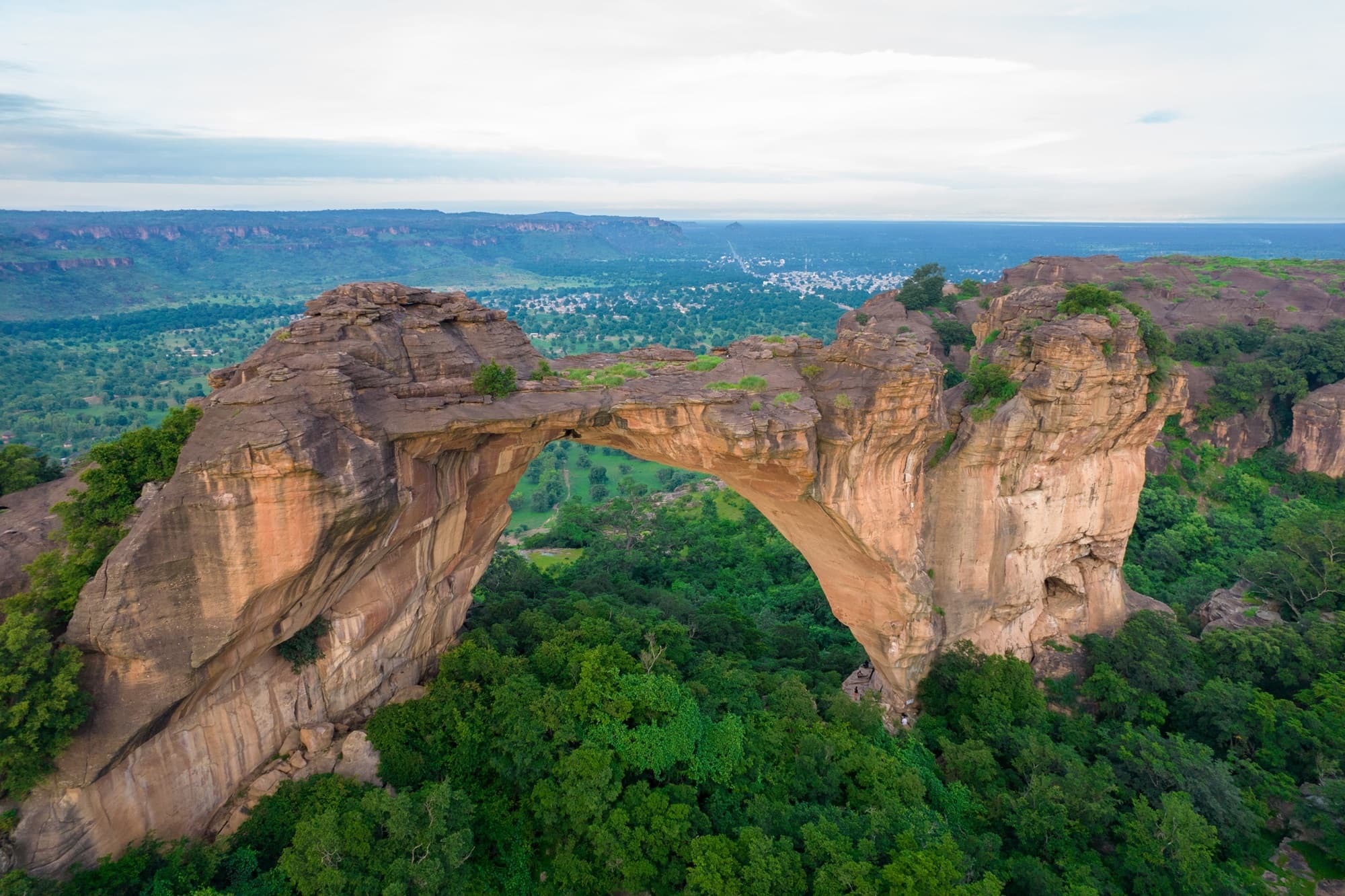 Aerial view of a large natural ochre sandstone arch on the Dogon escarpment, dense forest below and, beyond, the plain, a town and the line of the cliffs on the horizon. — API-Mali